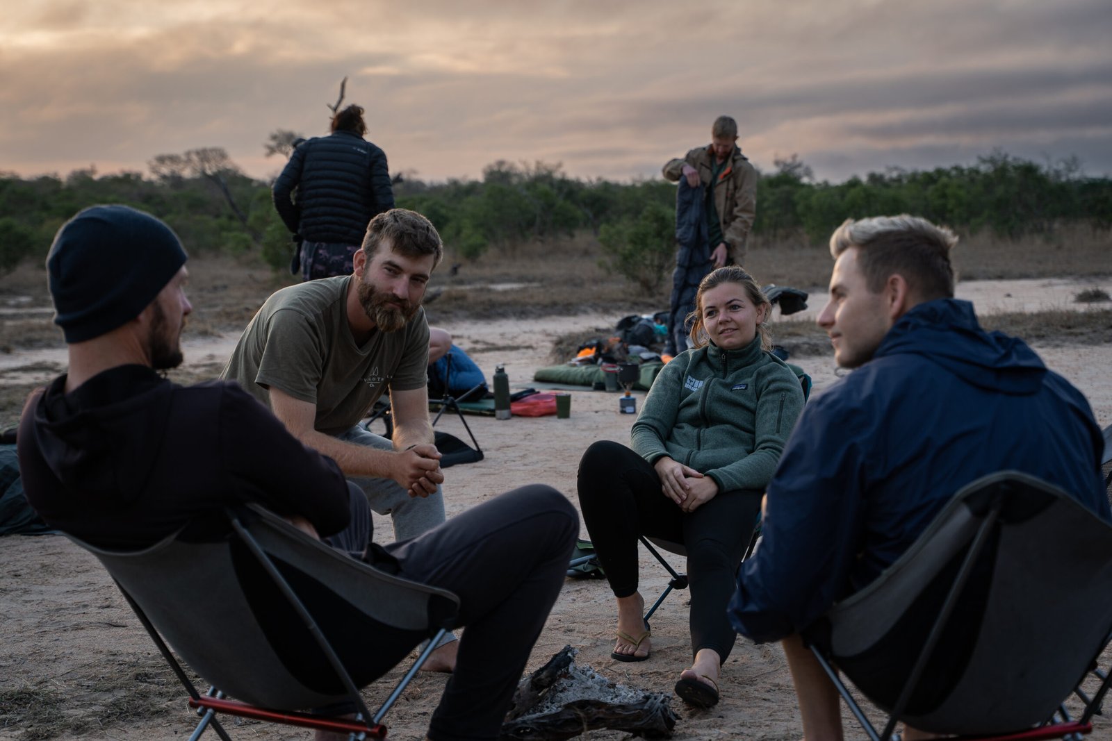 Vier deelnemers zitten op campingstoeltjes rond de gedoofde kampvuurplaats in het bushkamp tijdens de Primitive Trail in Greater Kruger, terwijl anderen hun slaapzakken opruimen.