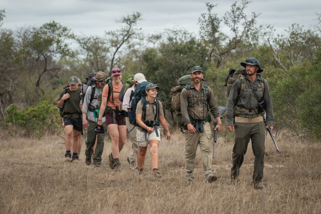 De gids loopt voorop tijdens de Primitive Trail wandelsafari in Greater Kruger, gevolgd door Daphne en de deelnemers in een rij met hun backpacks door het gras en tussen de bomen.