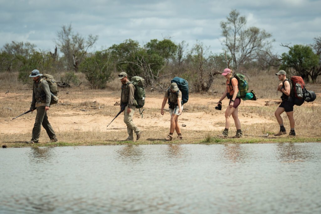 Daphne wijst naar iets aan de rand van een waterplas tijdens de Primitive Trail wandelsafari in Greater Kruger, terwijl de gidsen en andere deelnemers meekijken.