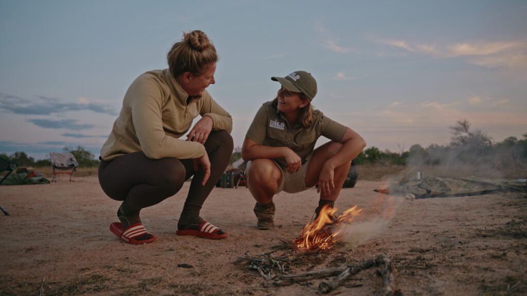 Daphne en deelnemer Simone kijken elkaar lachend aan bij het kampvuur dat ze net hebben gemaakt, met het bushkamp van de Primitive Trail op de achtergrond.