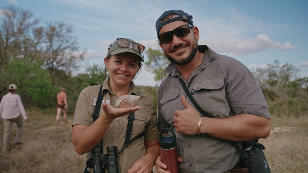 Daphne en deelnemer Ruan lachen naar de camera terwijl Daphne een gevonden huisje van een Afrikaanse reuzenslak laat zien tijdens de Primitive Trail in Greater Kruger. Op de achtergrond wandelen de andere deelnemers.